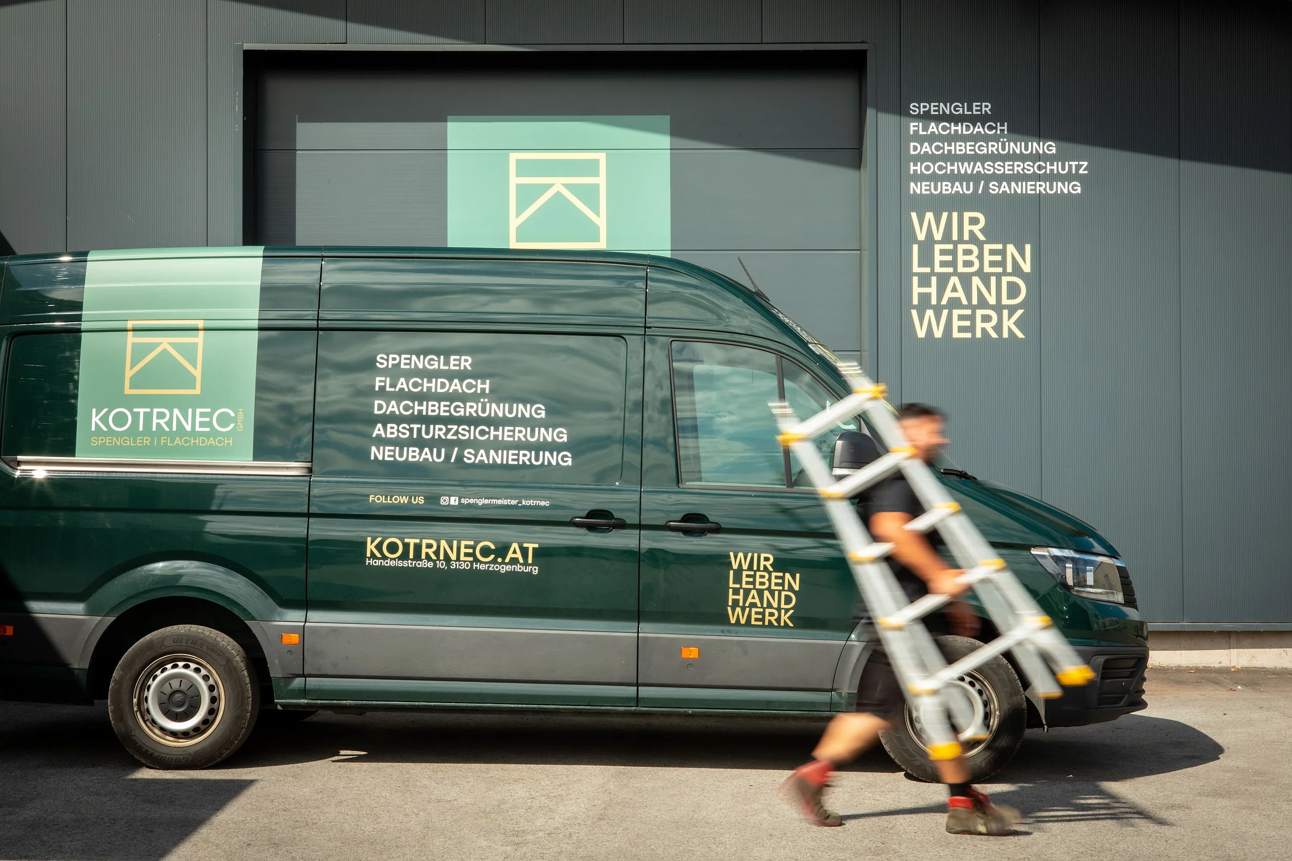 Firmenwagen mit Logo vor der Werkstatt. Davor geht ein Mitarbeiter mit einer Leiter.